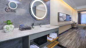 Un baño moderno con tocador de mármol, dos espejos redondos, un lavamanos y un espejo grande encima. Hay toallas y una planta en el tocador, y un televisor montado en la pared.
