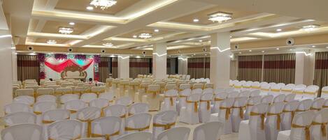 Une salle de banquet avec des rangées de chaises blanches ornées de rubans dorés, une scène avec un décor floral et un lustre.