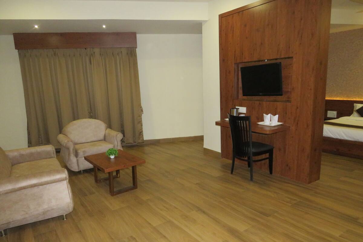 A hotel room with a wooden floor, a sofa, a small table with a plant, a TV mounted on a wooden panel, and a bed with a headboard.