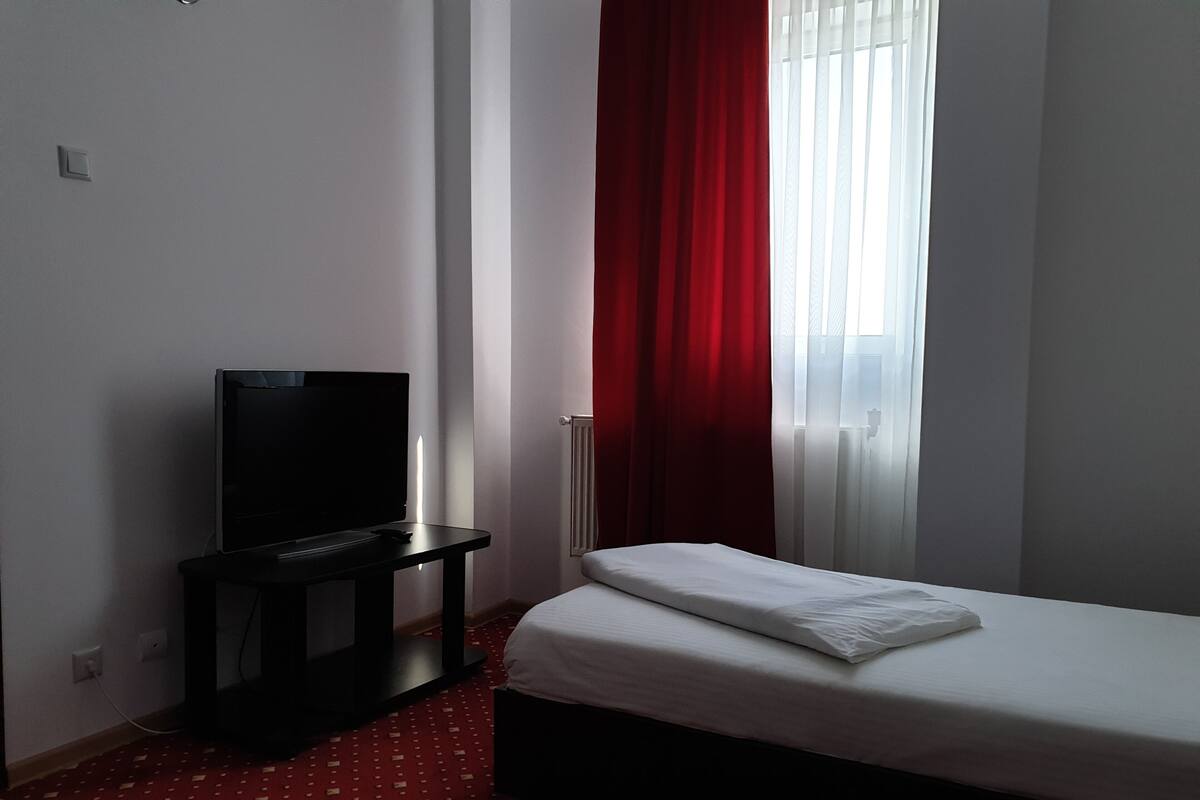 A hotel room with a bed, a TV on a stand, and a window with red curtains.