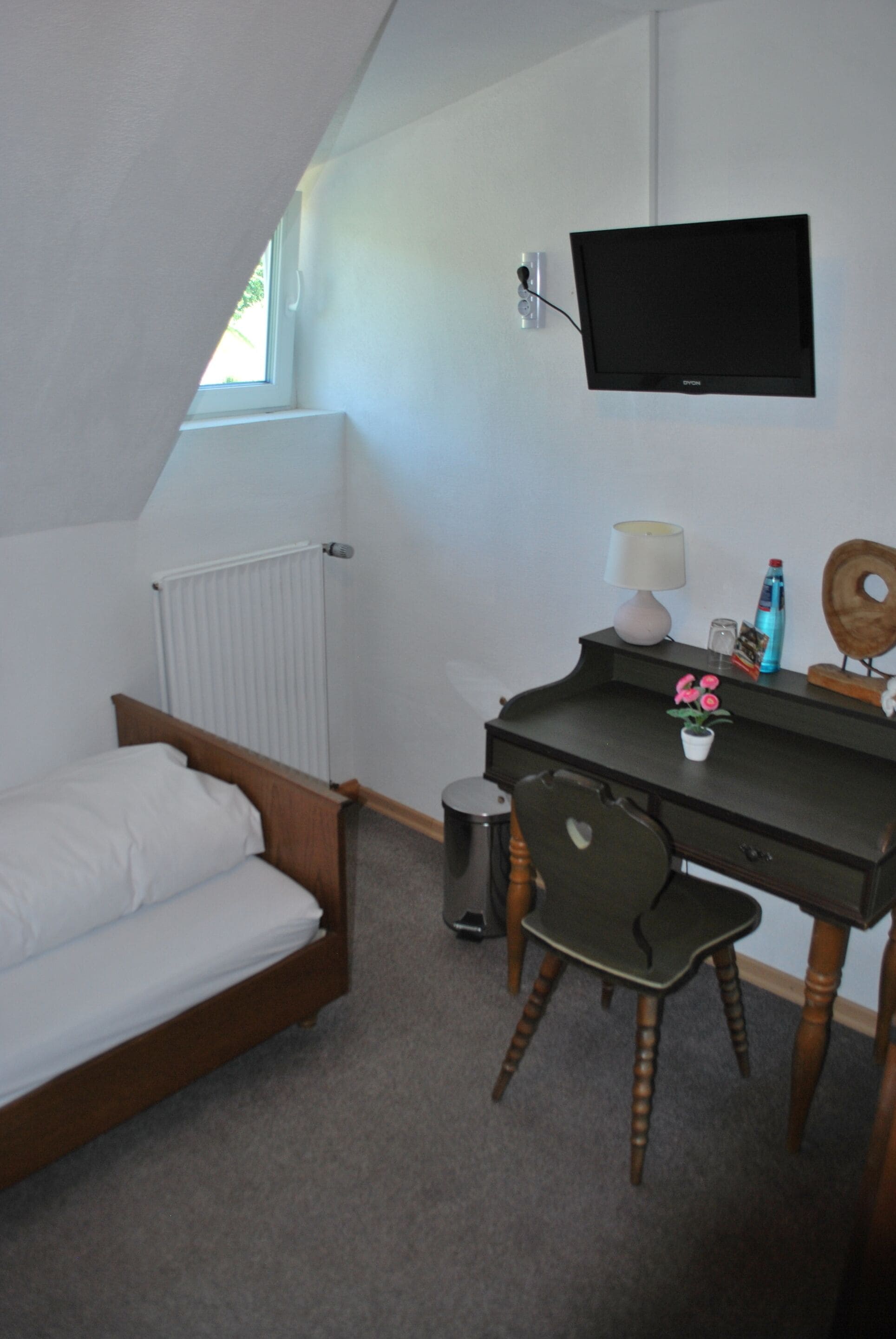A bedroom with a bed, a desk with a chair, a TV mounted on the wall, and a window.