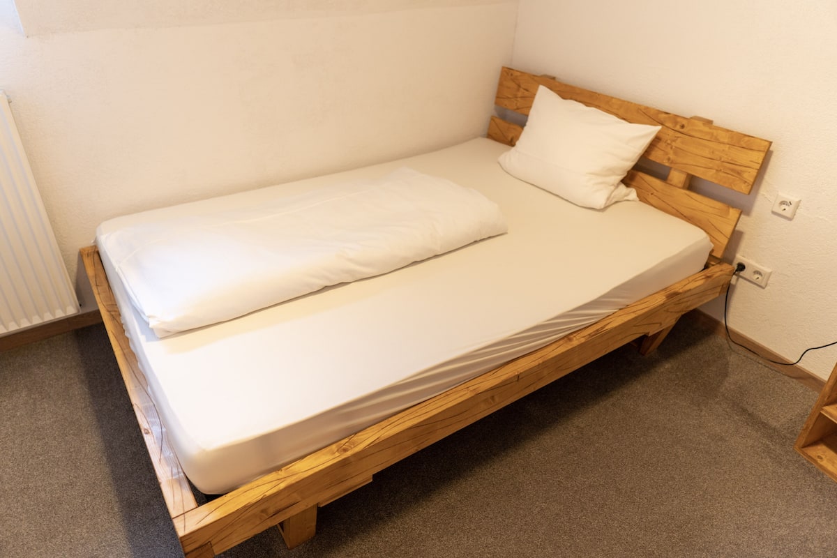 A single bed with a wooden frame and white bedding in a room with a carpeted floor and white walls.