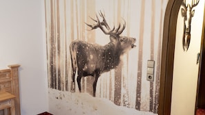 Huoneessa on suuri seinämaalaus, jossa on kuvattuna peura, punakuvioinen matto, puinen lipasto ja ovi, jossa on peuranpääaiheinen koriste.