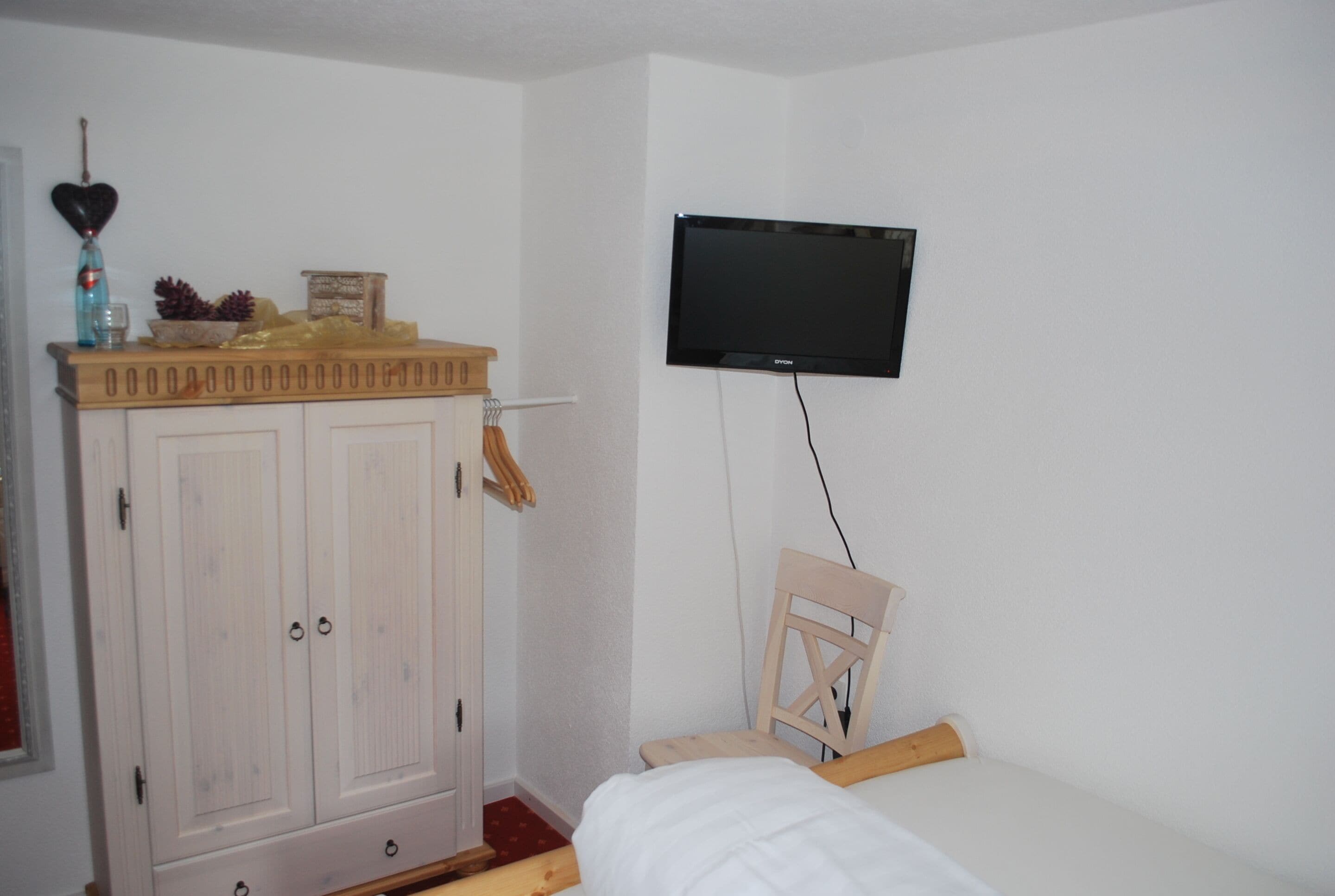 Quarto com cama, guarda-roupa de madeira, televisão na parede e uma cadeira de madeira pequena.