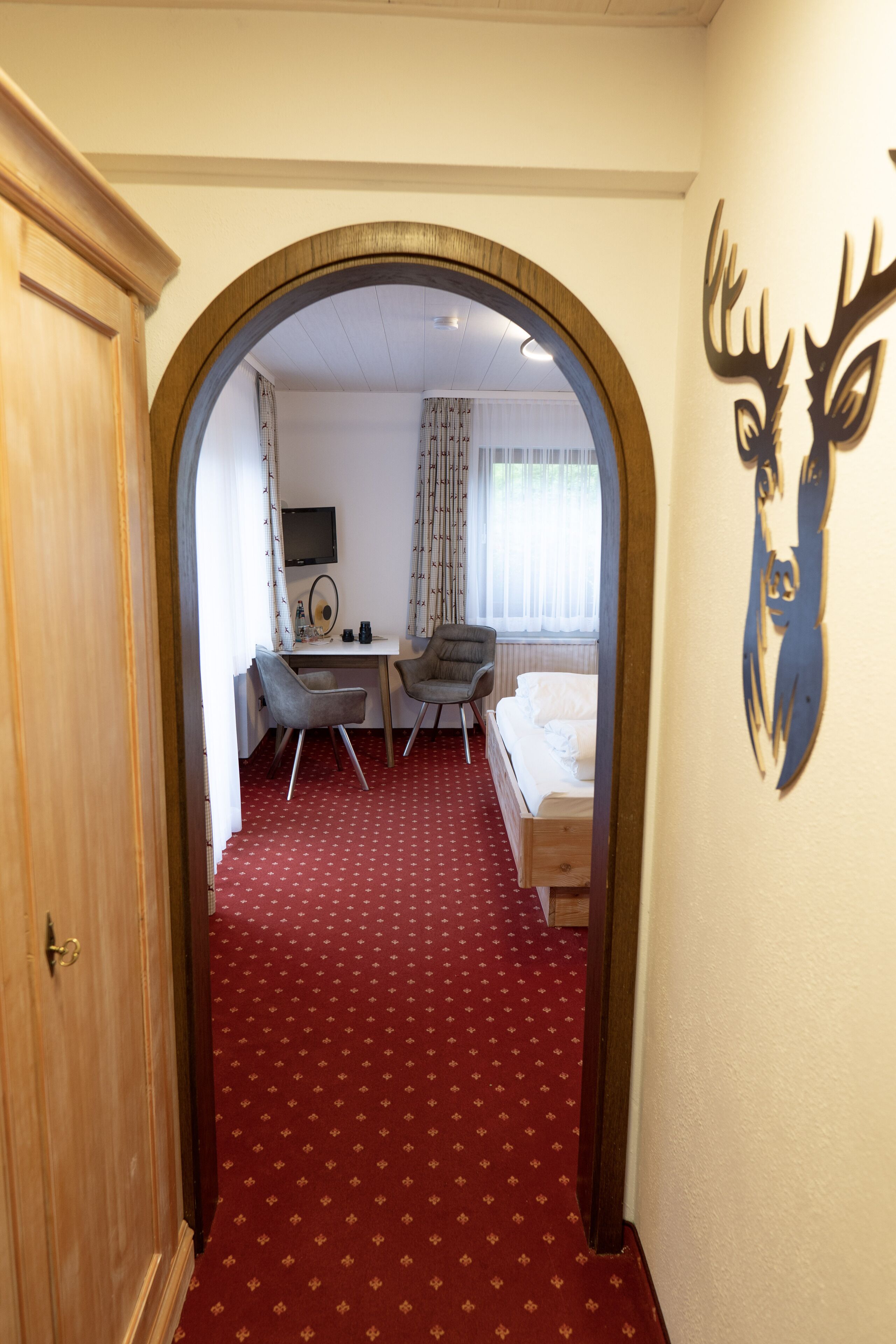A hotel room with a red carpet, a desk with a chair, a bed, and a TV on the wall.