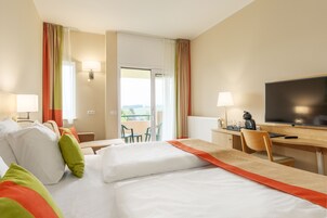 A hotel room with two beds, a desk with a TV, a chair, and a balcony with a view.
