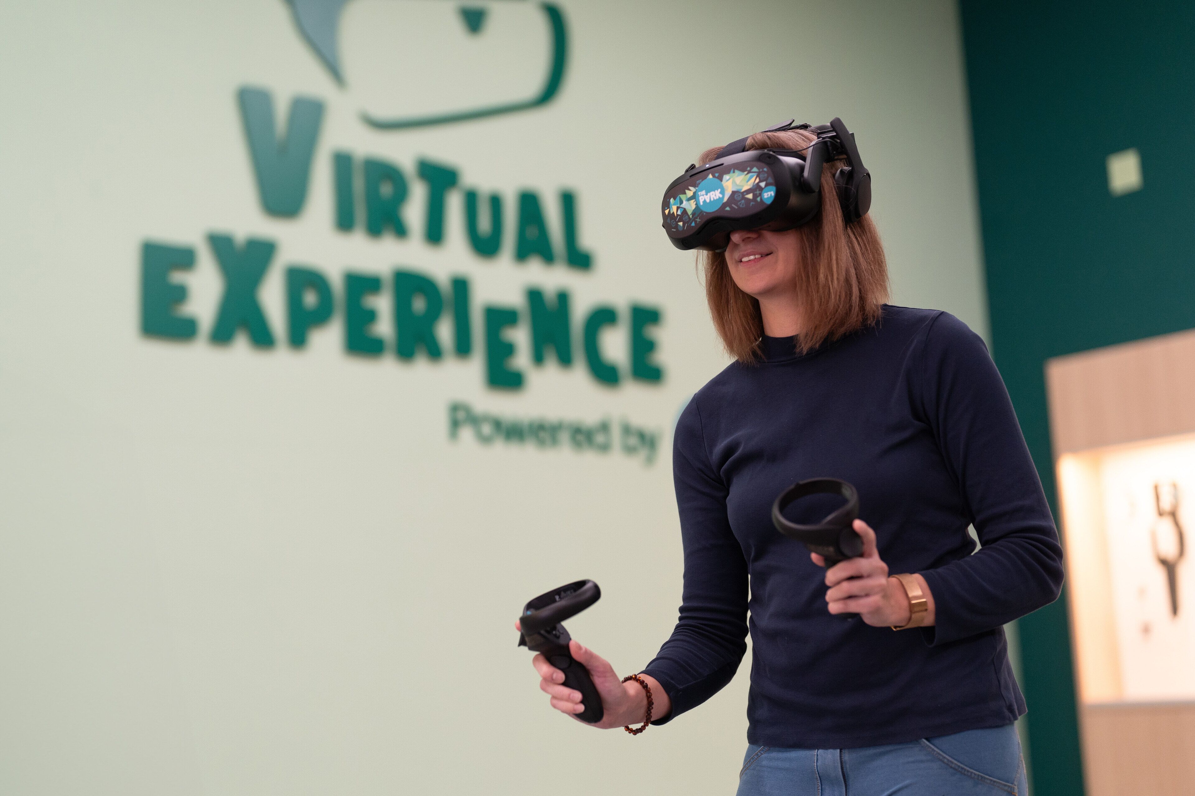 Una persona con un casco de realidad virtual, sosteniendo un control de RV, de pie frente a un letrero que dice 