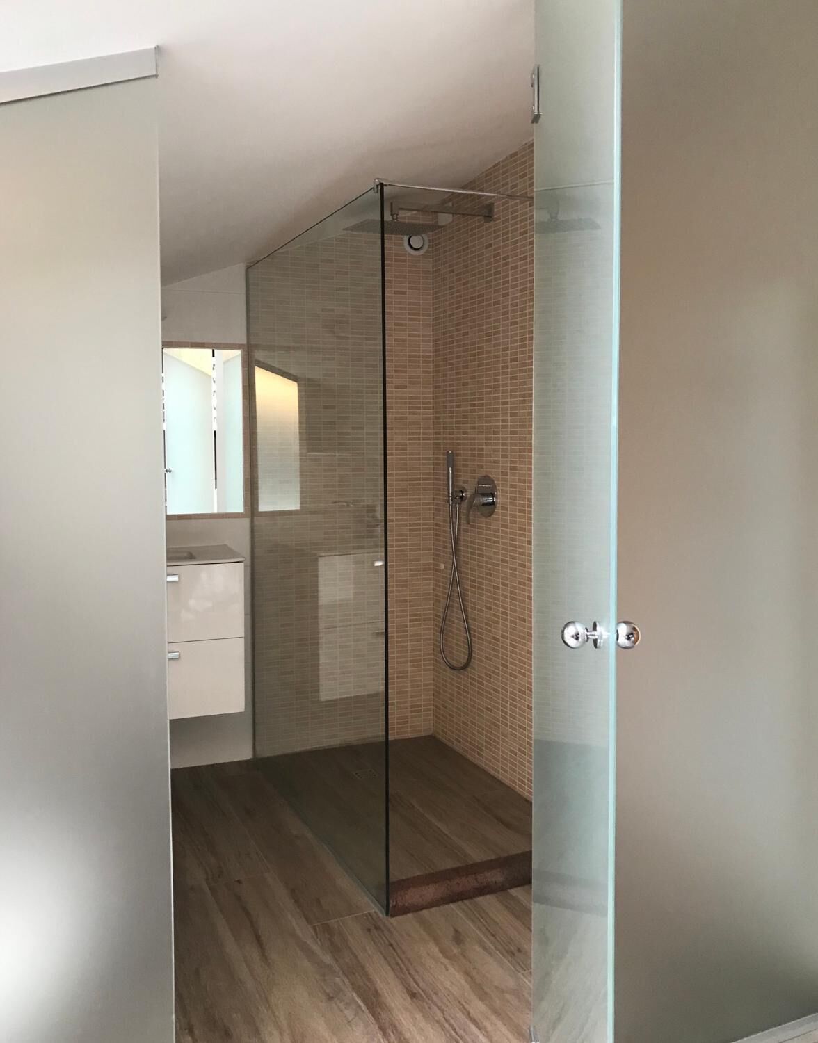 Et moderne badeværelse med glasvæg omkring brusebad, trægulv og et synligt område med håndvask.