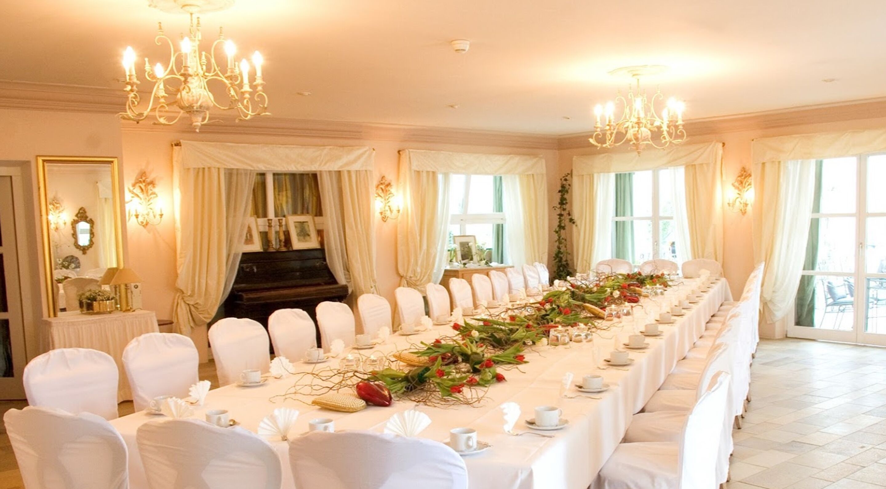 A long banquet table with white chairs, chandeliers, and draped curtains.