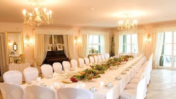 Uma longa mesa de banquete com cadeiras brancas, lustres e cortinas penduradas.