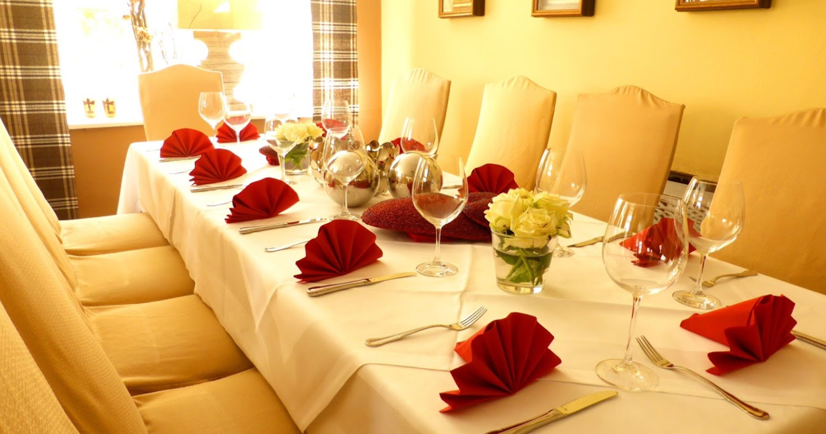 A dining table set with red napkins, wine glasses, and cutlery, with a vase of flowers in the center.