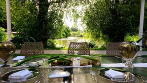 Una mesa para comer al aire libre dispuesta con servilletas blancas, cubiertos y cristalería, rodeada de sillas de mimbre, con vista a un estanque y abundante vegetación.