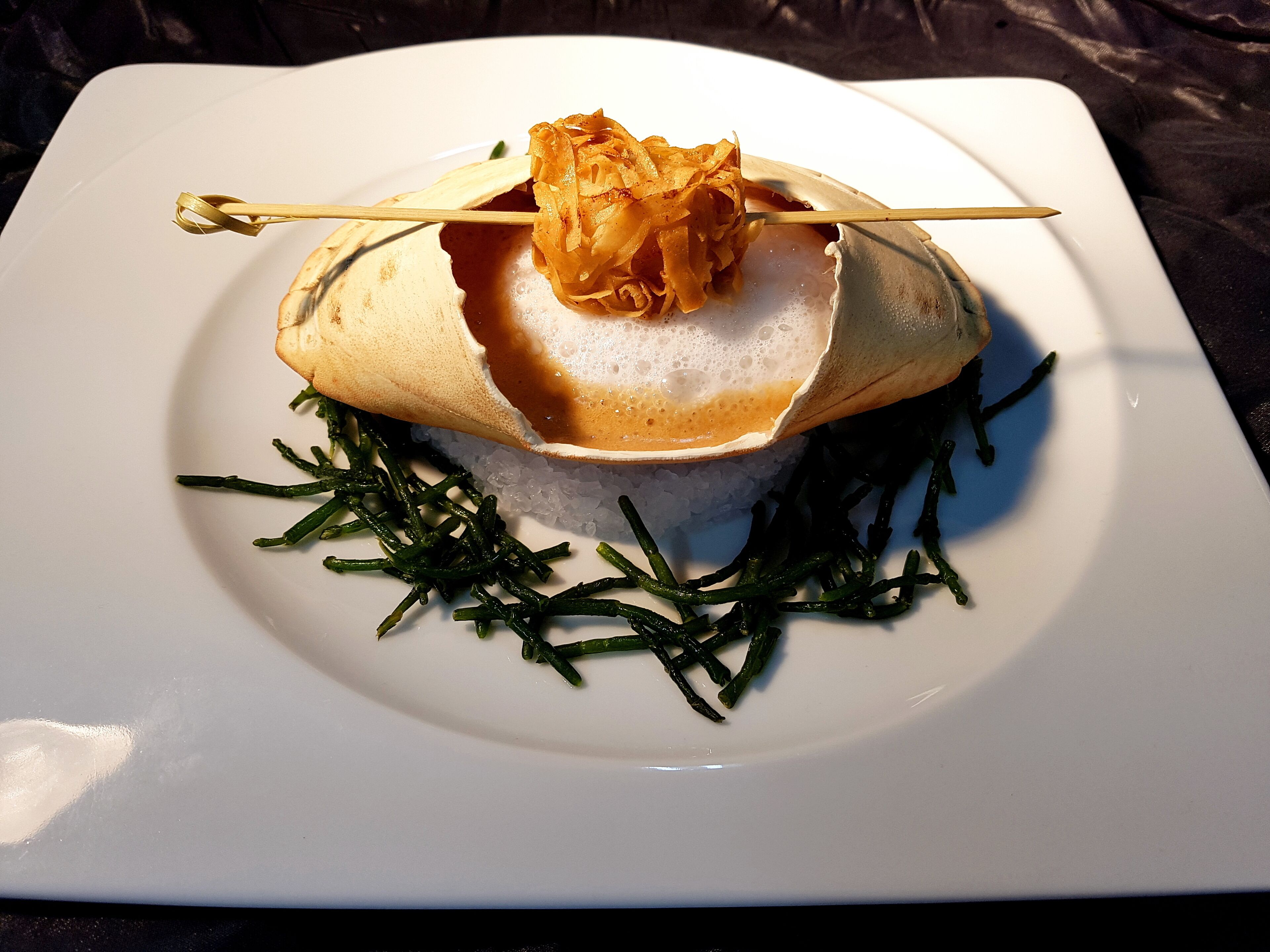 A white plate with a dish consisting of rice, a piece of seaweed, and a crispy topping on a skewer.