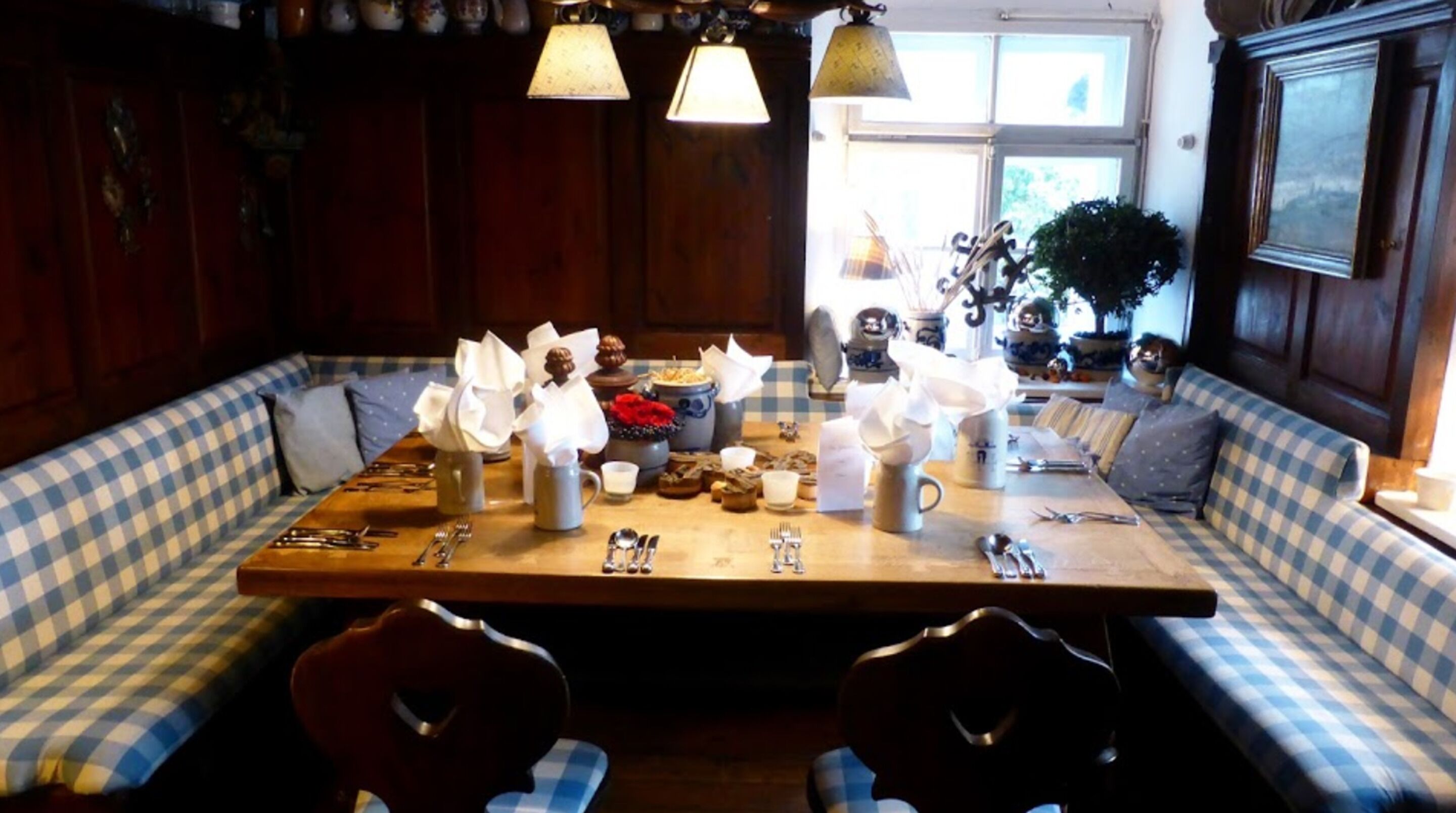Un comedor con banco de asiento a cuadros, una mesa de madera dispuesta para cuatro personas y elementos decorativos como un jarrón con flores y un reloj.