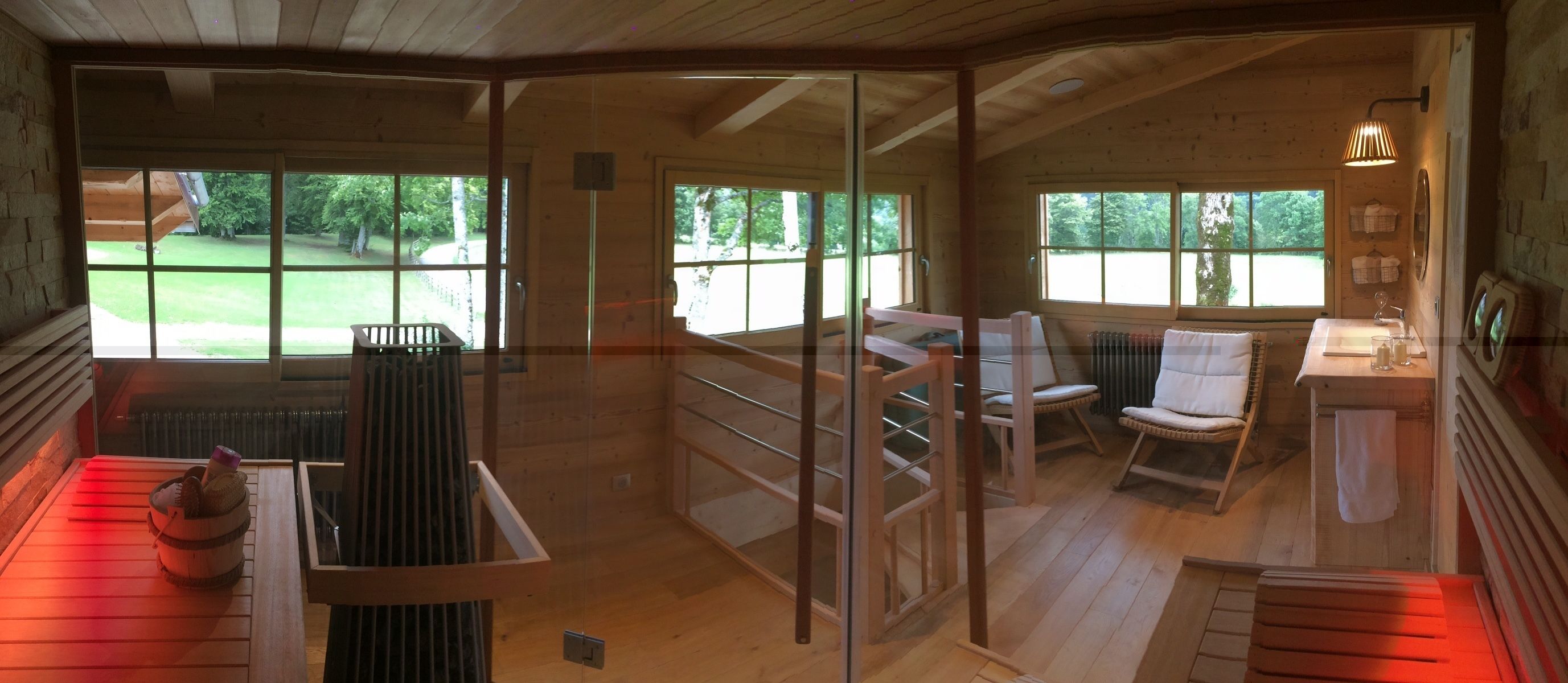 Una cabaña de madera con puerta de cristal que da a un balcón, zona de estar con sillas y vistas a un paisaje verde.