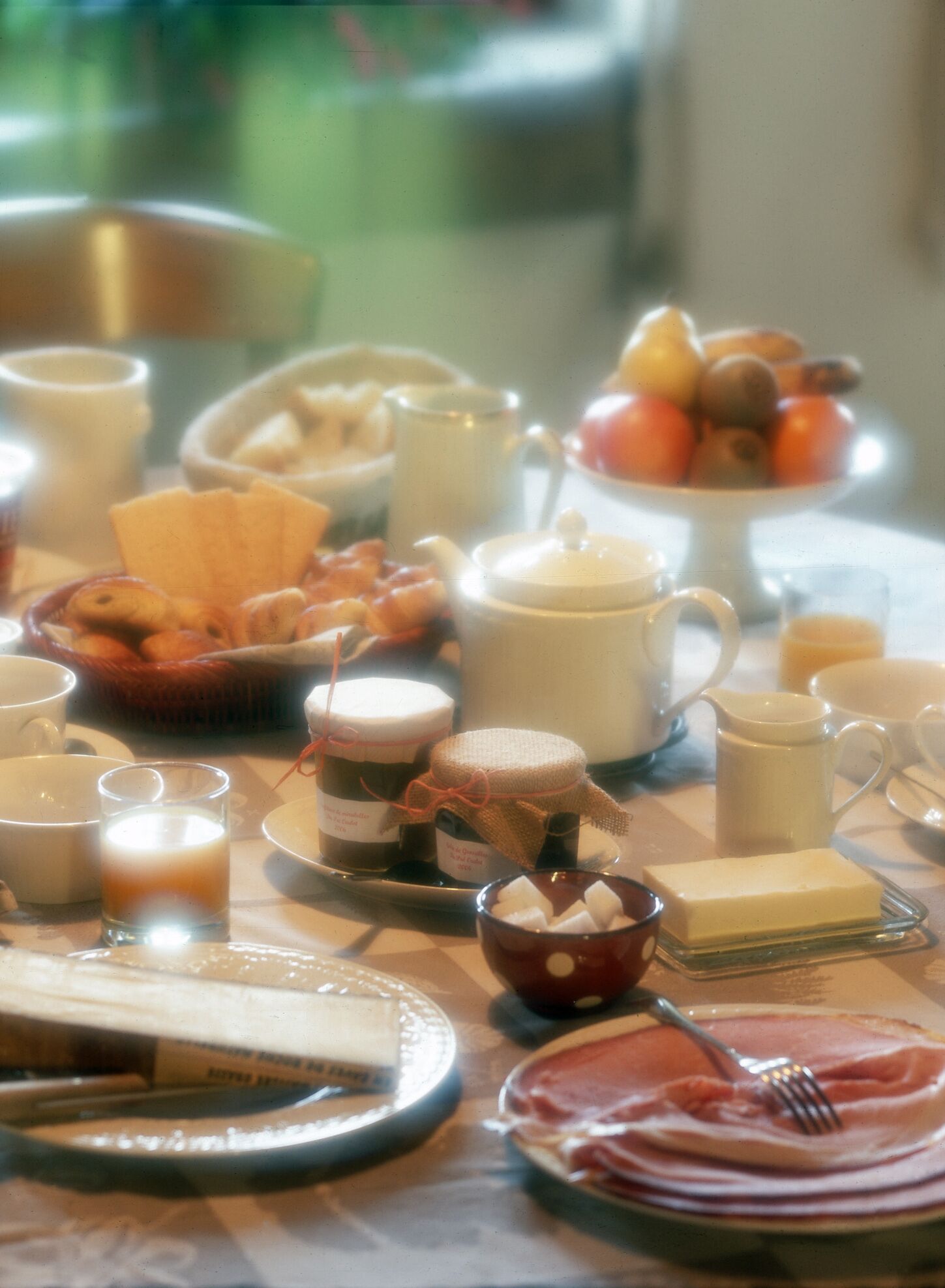A breakfast table with various food items and drinks, including a plate of ham, a bowl of fruit, and several cups and mugs.