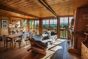 Interior de una cabaña de madera con comedor, sofá y ventanas amplias que ofrecen vistas al exterior.