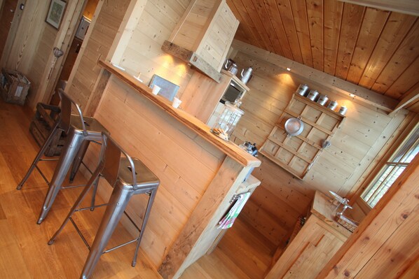 Interior de madera con una cocina que incluye una barra, taburetes y diferentes electrodomésticos.