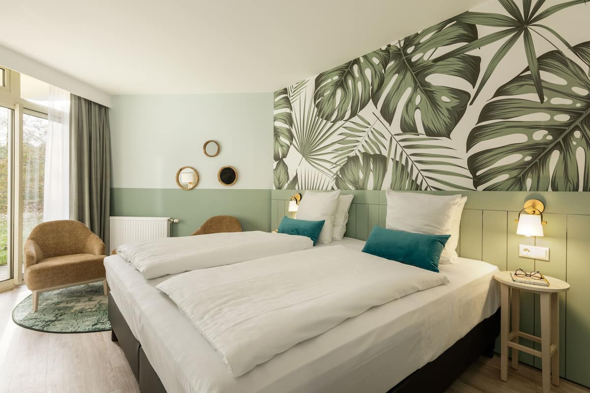 A hotel room with two beds, a round rug, a chair, a wall with a tropical leaf pattern, and a nightstand with a lamp.