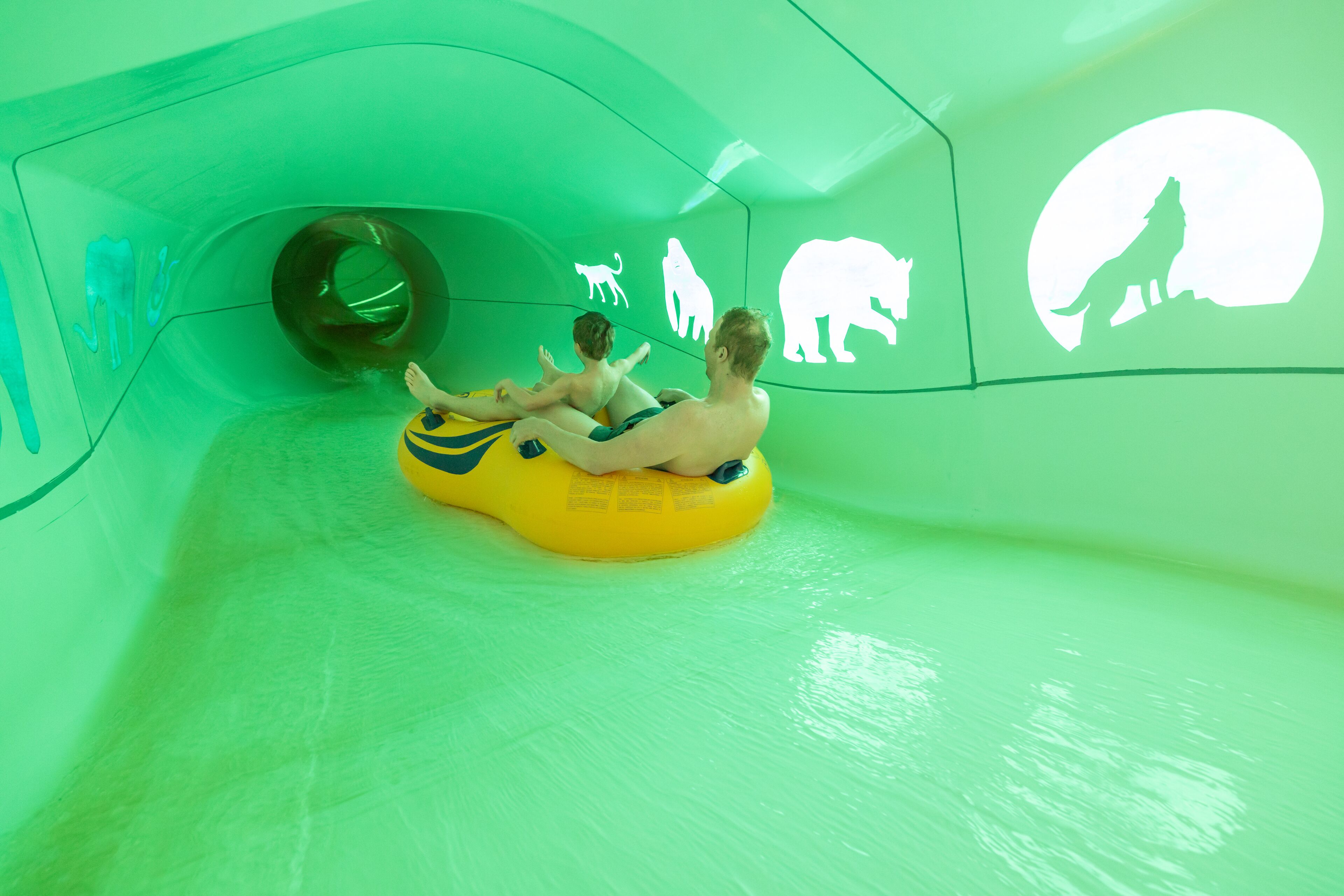 Two people sliding on a yellow tube in a green-themed water slide area with animal silhouettes on the walls.