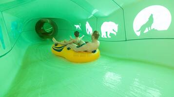 Two people sliding on a yellow tube in a green-themed water slide area with animal silhouettes on the walls.