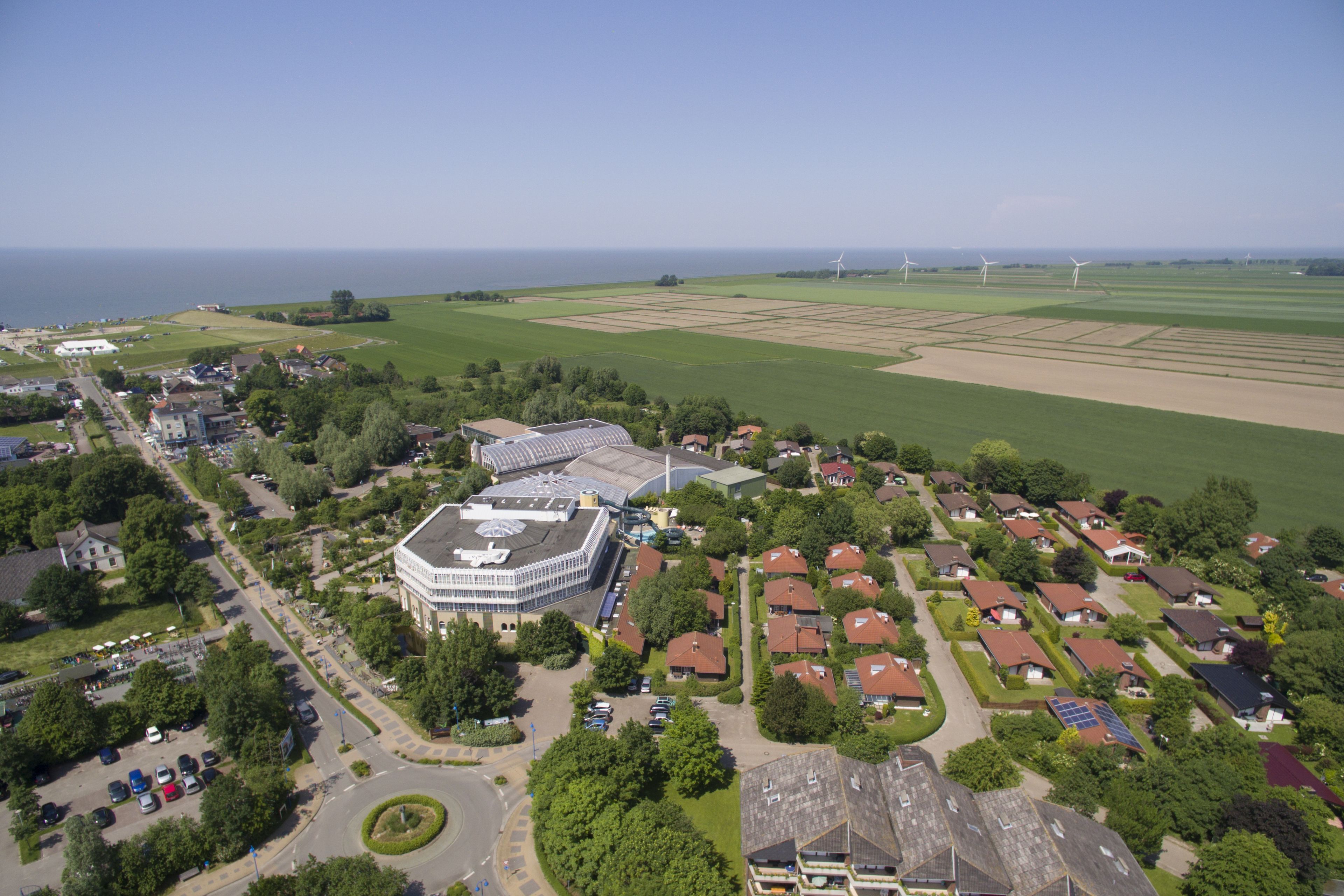 Vista aérea de una zona residencial con casas, un edificio central con un techo distintivo y aerogeneradores a lo lejos.