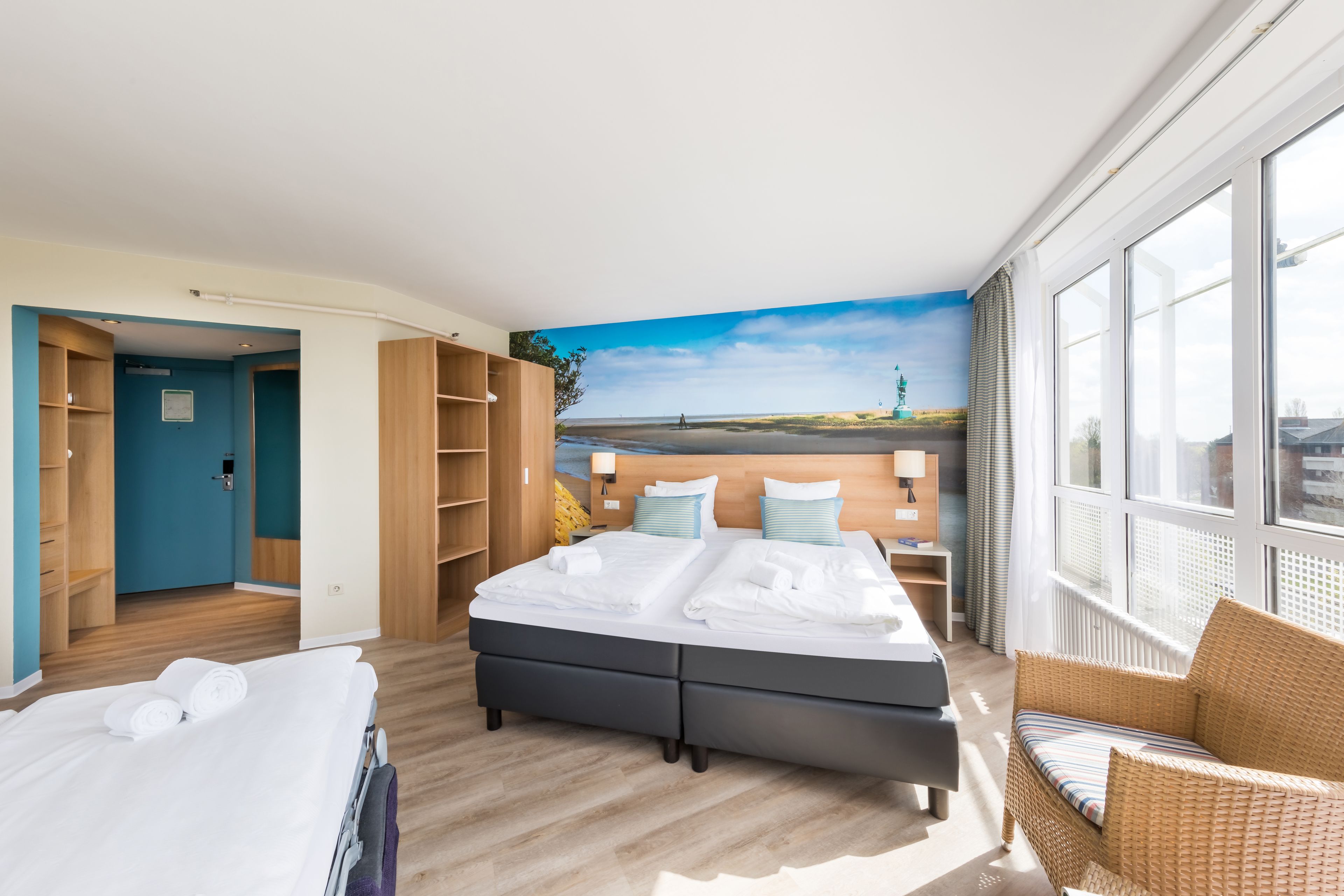 A hotel room with a large bed, a wicker chair, a view of the beach, and a wardrobe.