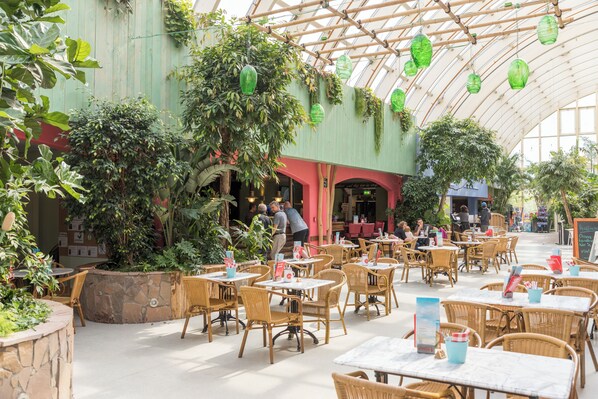 An indoor cafe with wicker tables and chairs, stone planters, and hanging green lanterns.