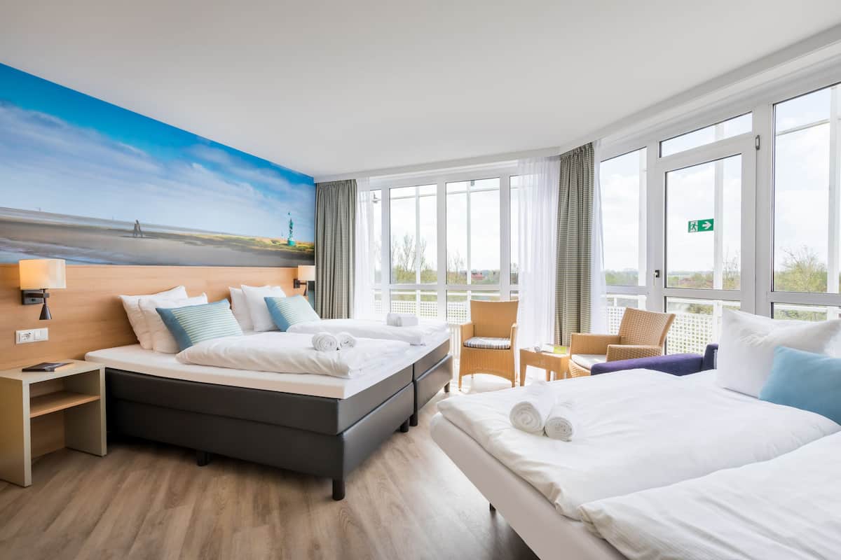 Hotelkamer met een groot bed, een kleiner bed, een bureau en een stoel. Er hangt een schilderij aan de muur en er is een raam met gordijnen.