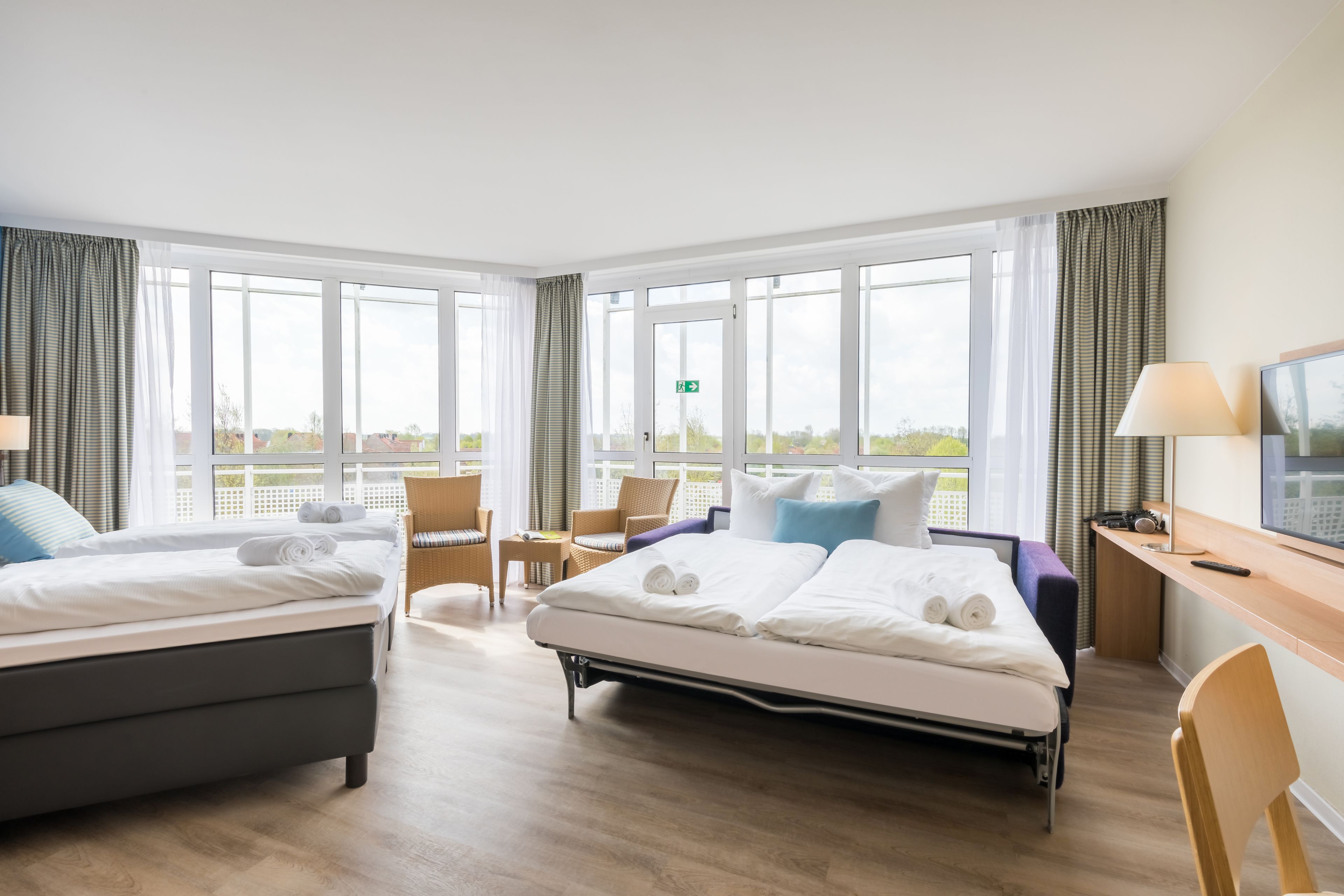 A hotel room with two beds, a desk with a lamp, a chair, and large windows with curtains.