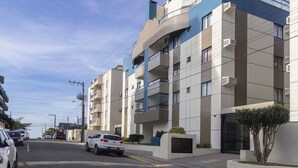 A multi-story residential building with balconies, a gated entrance, and a parking area with cars. - Cayman 123 - Apto 80M Praia de Bombas (Bombinhas)