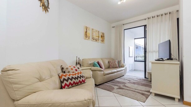 A living room with two beige sofas, a television on a stand, and a view through a glass door to the outside. - Cayman 123 - Apto 80M Praia de Bombas (Bombinhas)