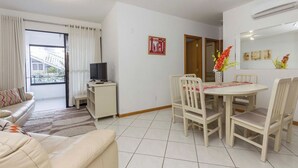 A modern living room with a sofa, a dining table with chairs, a TV, and a window with curtains. - Cayman 123 - Apto 80M Praia de Bombas (Bombinhas)
