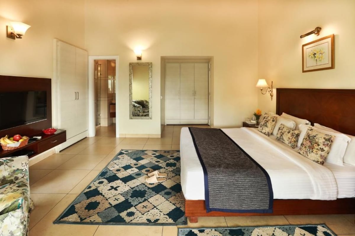 Habitación de hotel con cama, televisión, escritorio con cesta de fruta, espejo y puerta que conduce a otra habitación.