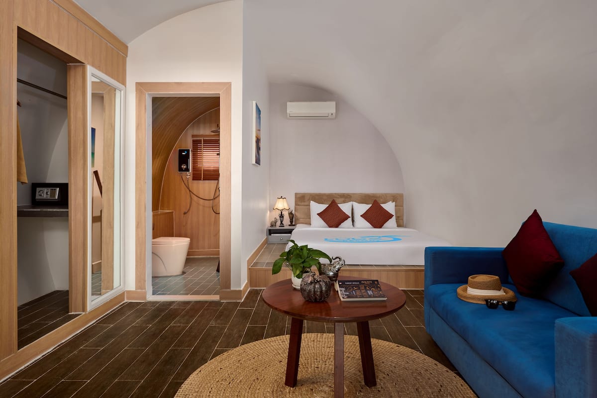 A bedroom with a bed, a blue sofa, a wooden wardrobe, a bathroom with a toilet, and a small table with a plant.