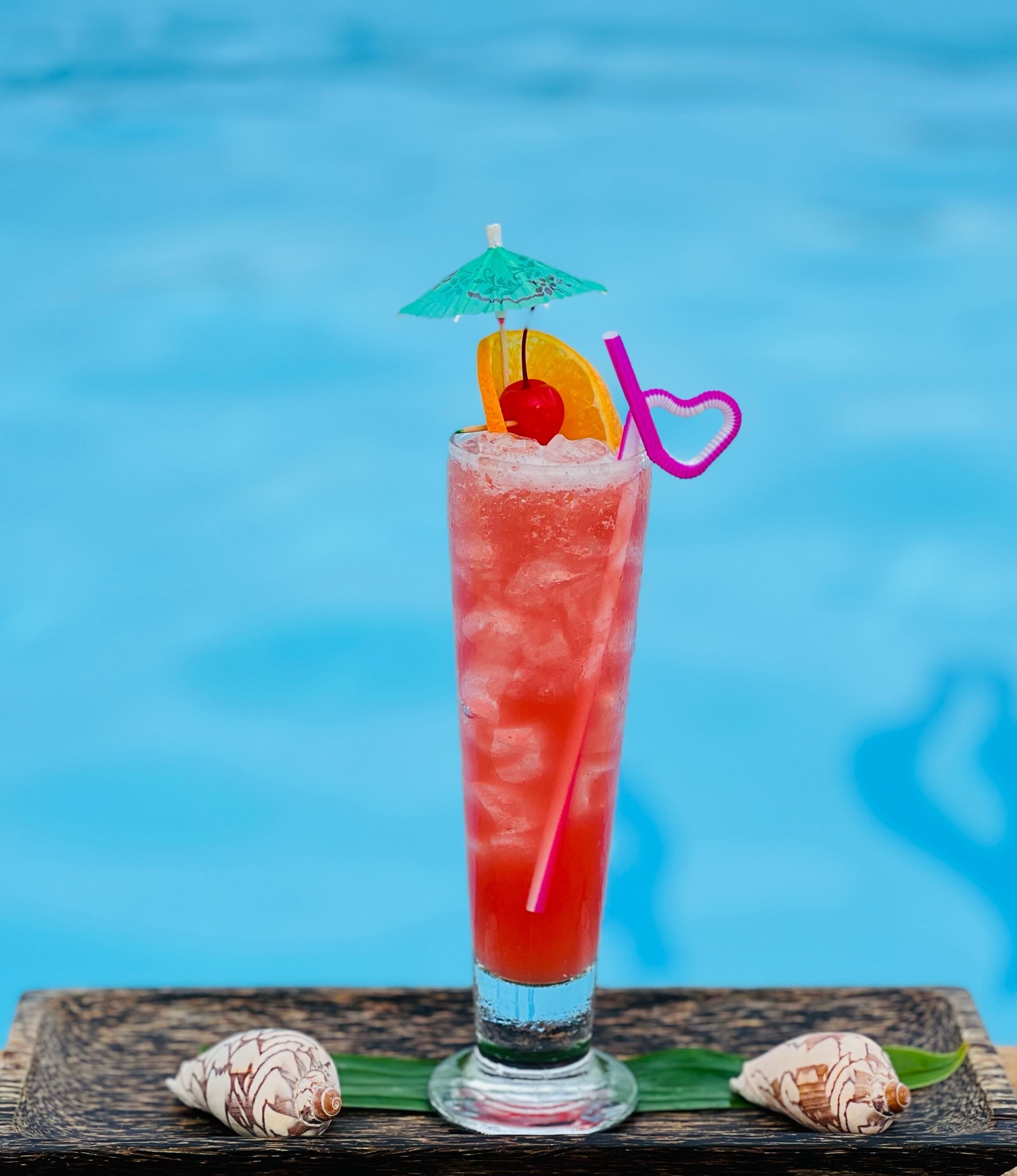Un grand verre contenant une boisson rose, une cerise, une tranche d’orange et une paille en forme de cœur, le tout sur un fond bleu.
