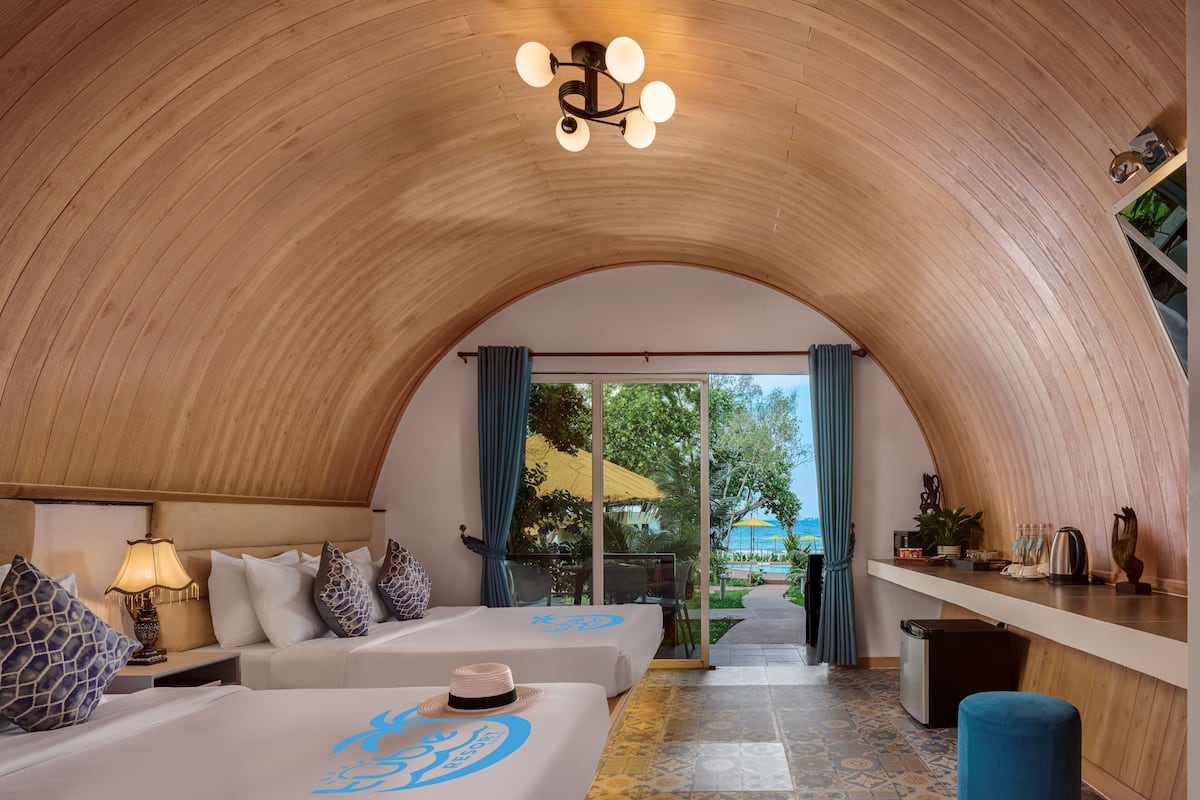 A room with a curved wooden ceiling, two beds with matching headboards, a small table with a lamp, and a view through a glass door to an outdoor area.
