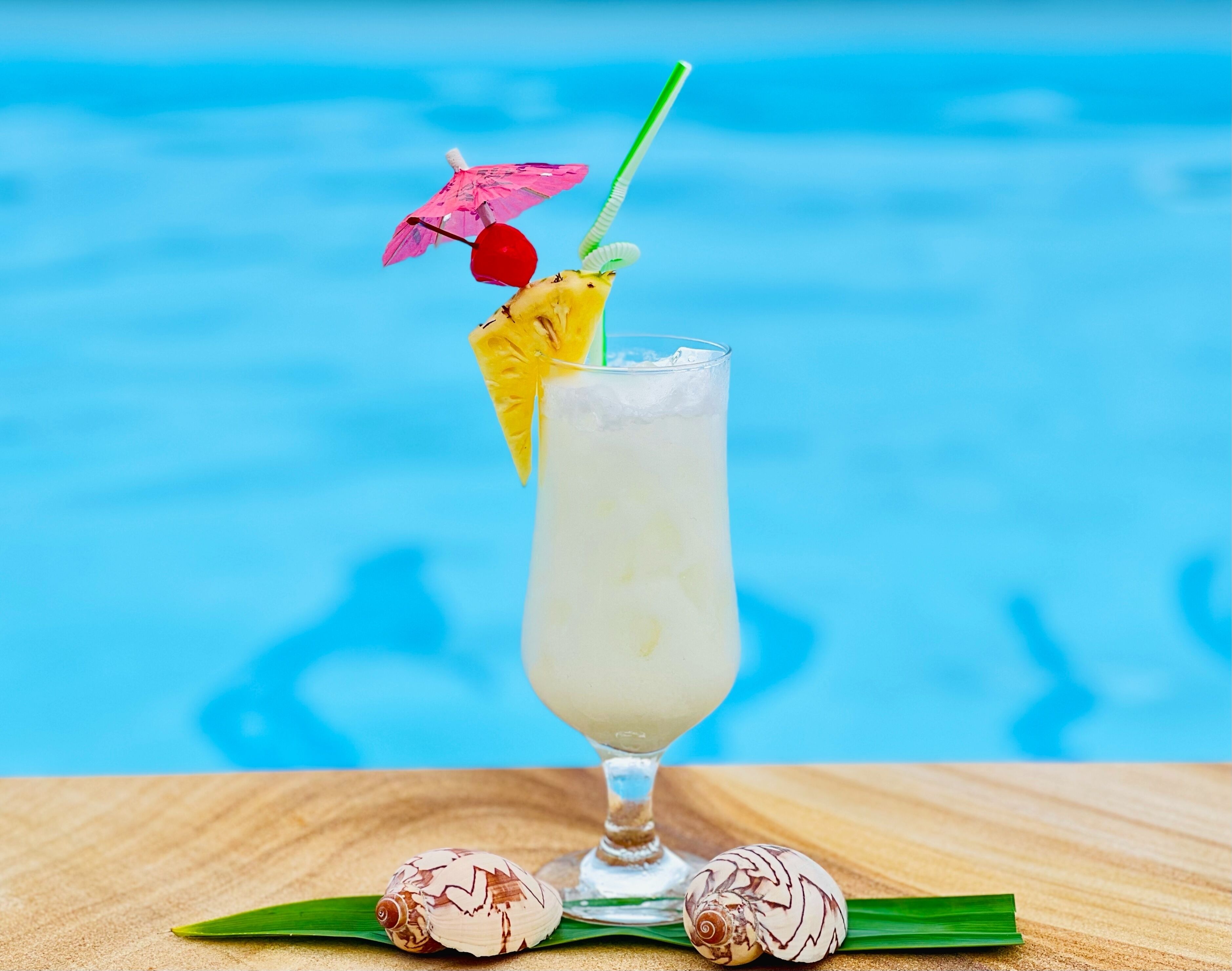 A cocktail with a straw, a cherry, and a decorative umbrella, placed on a wooden surface with a pool in the background.