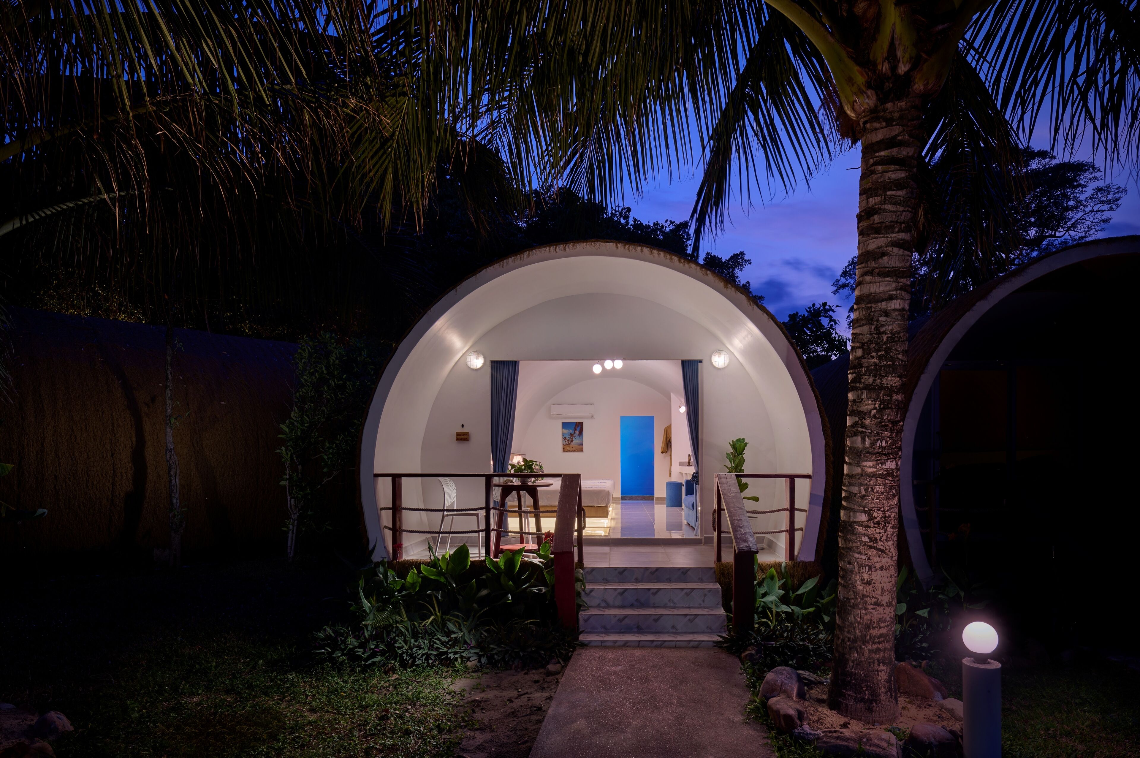 A dome-shaped building with a staircase leading to a door, surrounded by palm trees and plants.