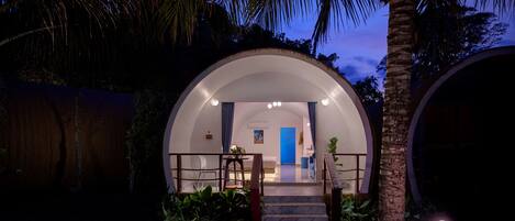 A dome-shaped building with a staircase leading to a door, surrounded by palm trees and plants.