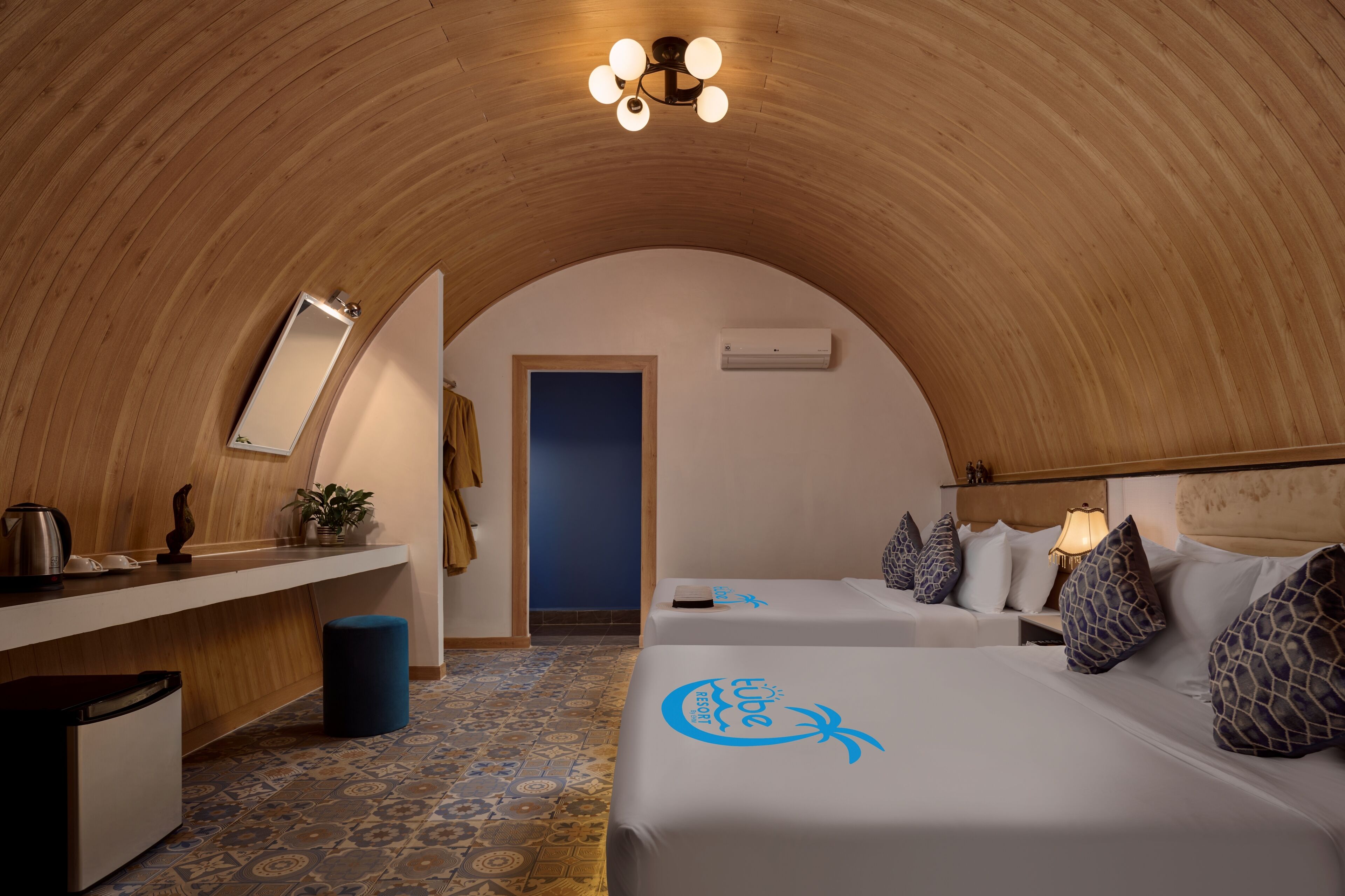 Une pièce dotée d’un plafond en bois aux lignes courbes, d’un bureau avec un miroir, de deux lits avec des oreillers assortis, d’une table de chevet avec une lampe et d’un sol à motifs.