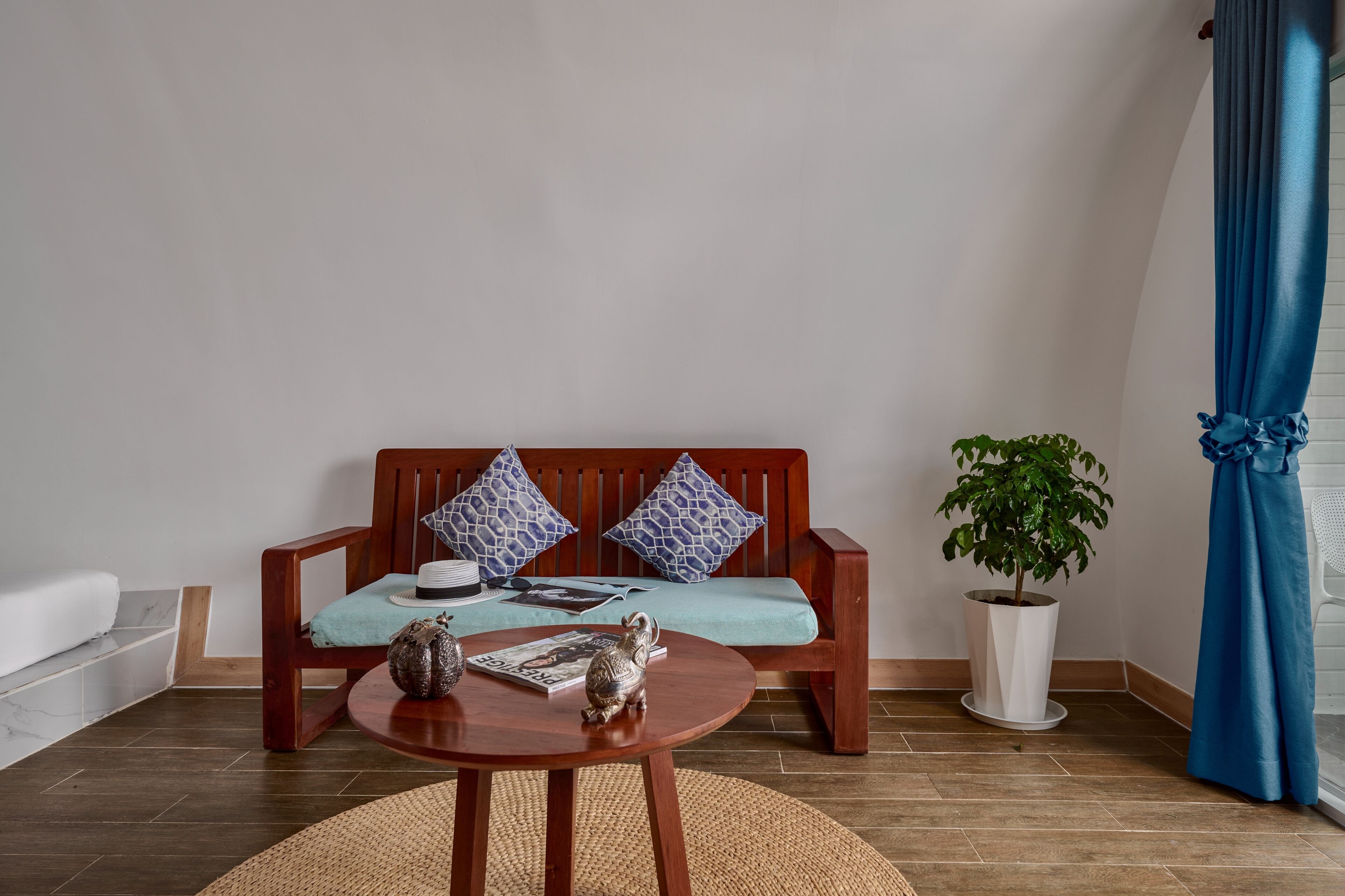 A wooden sofa with patterned cushions, a round wooden table with decorative items, a potted plant, and a blue curtain.