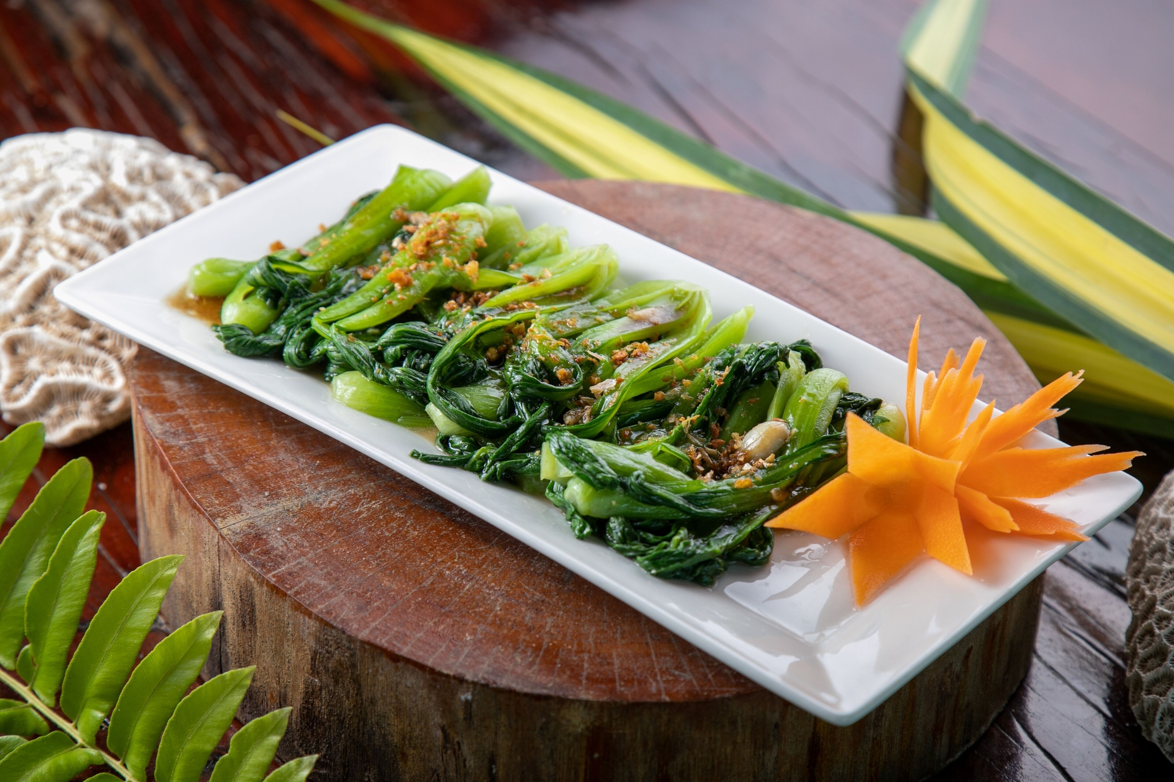 A rectangular white plate with a dish of stir-fried greens and a decorative orange garnish on a wooden table.