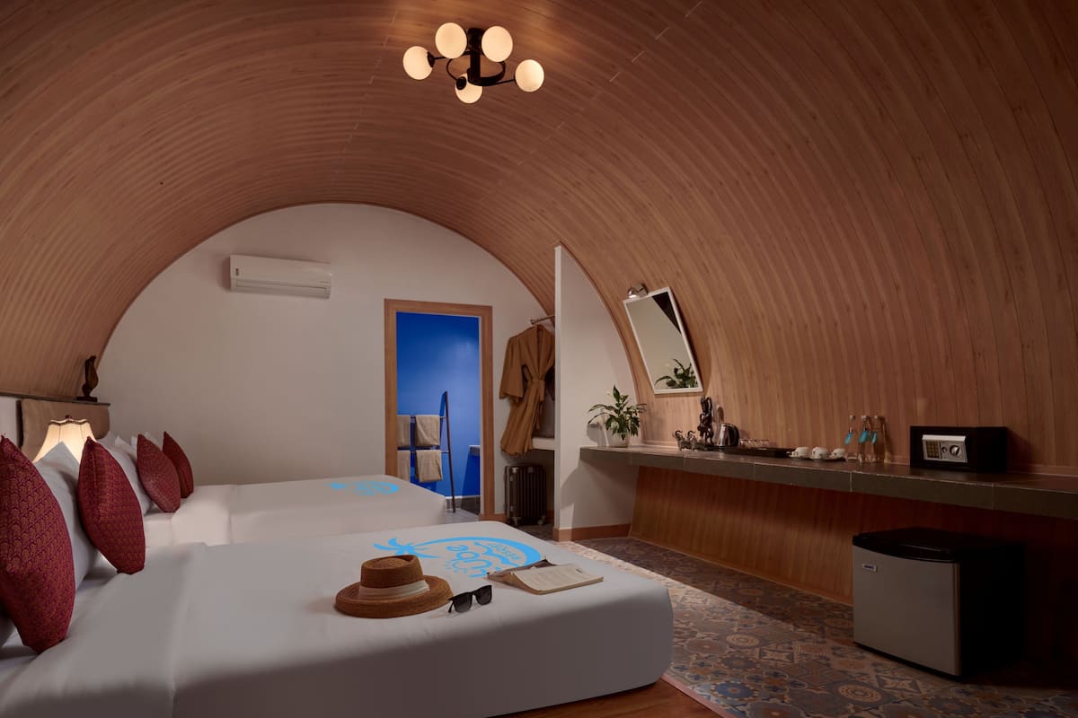 A cozy, curved-roofed room with a bed, a vanity with a mirror, and a small refrigerator.