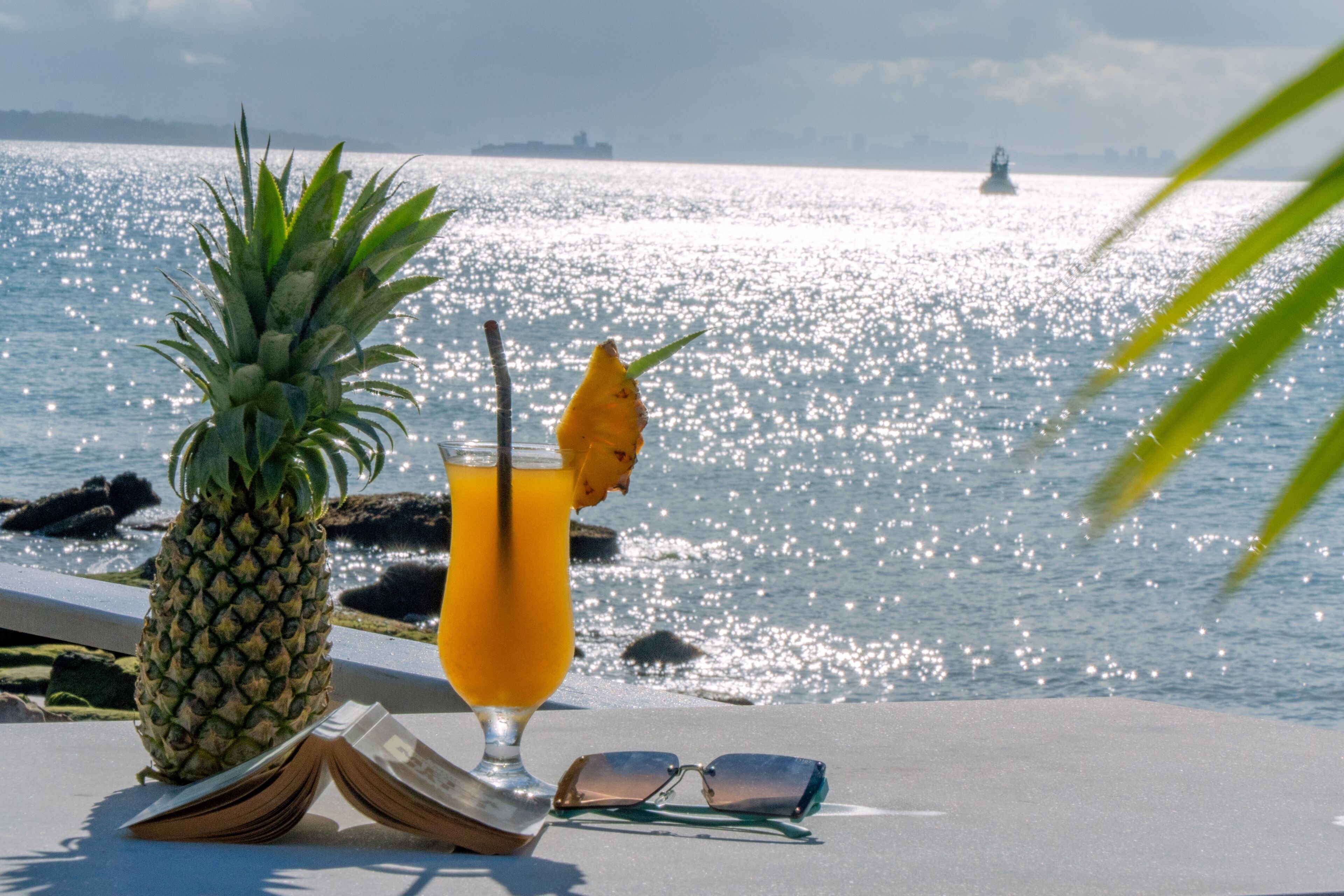 A tropical setting with a pineapple, a drink, and sunglasses on a table by the water.