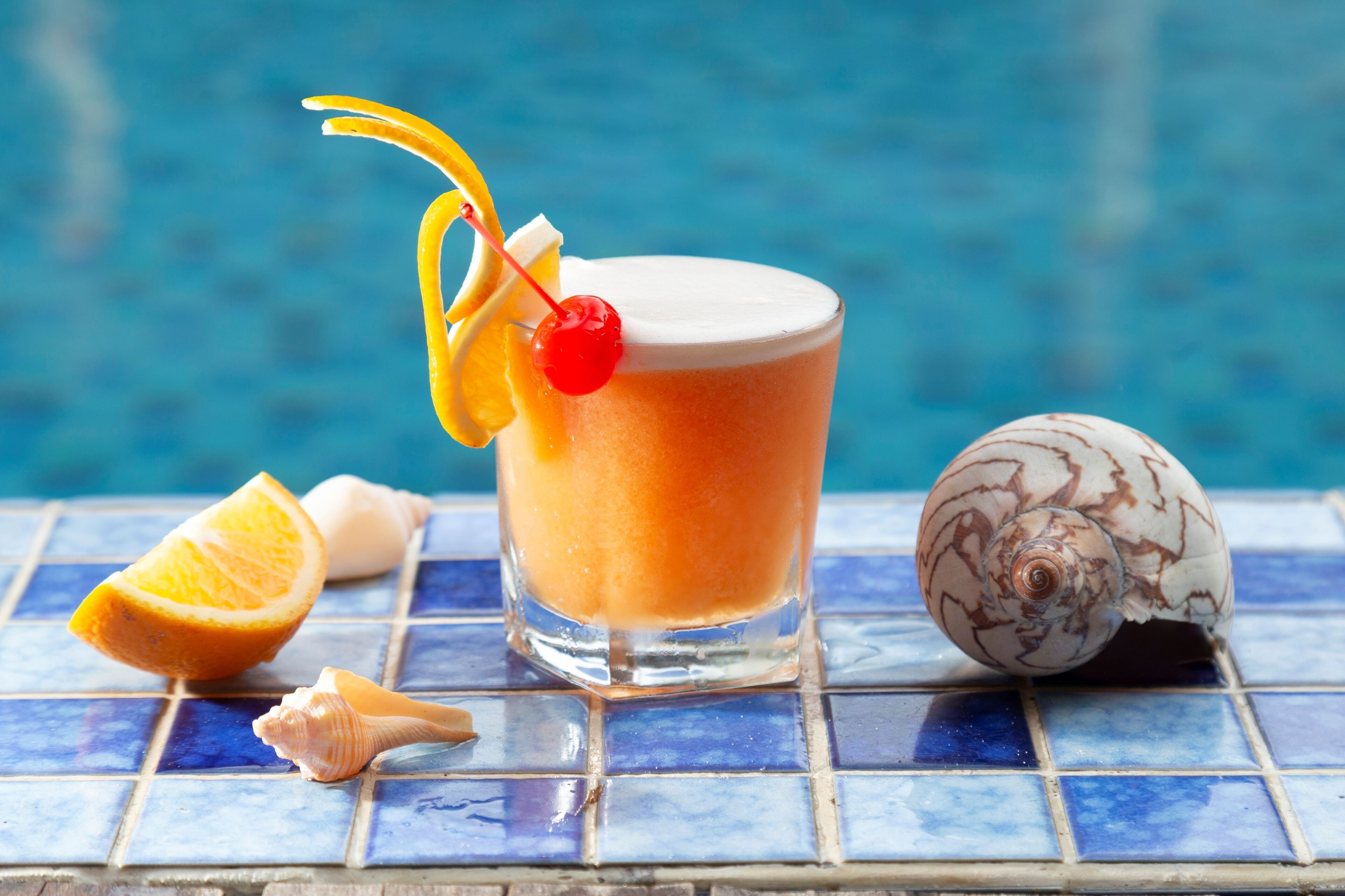 Een glas oranje drank met een kers, omringd door een helft van een sinaasappel, schelpen en een decoratieve schelp bij een zwembad.