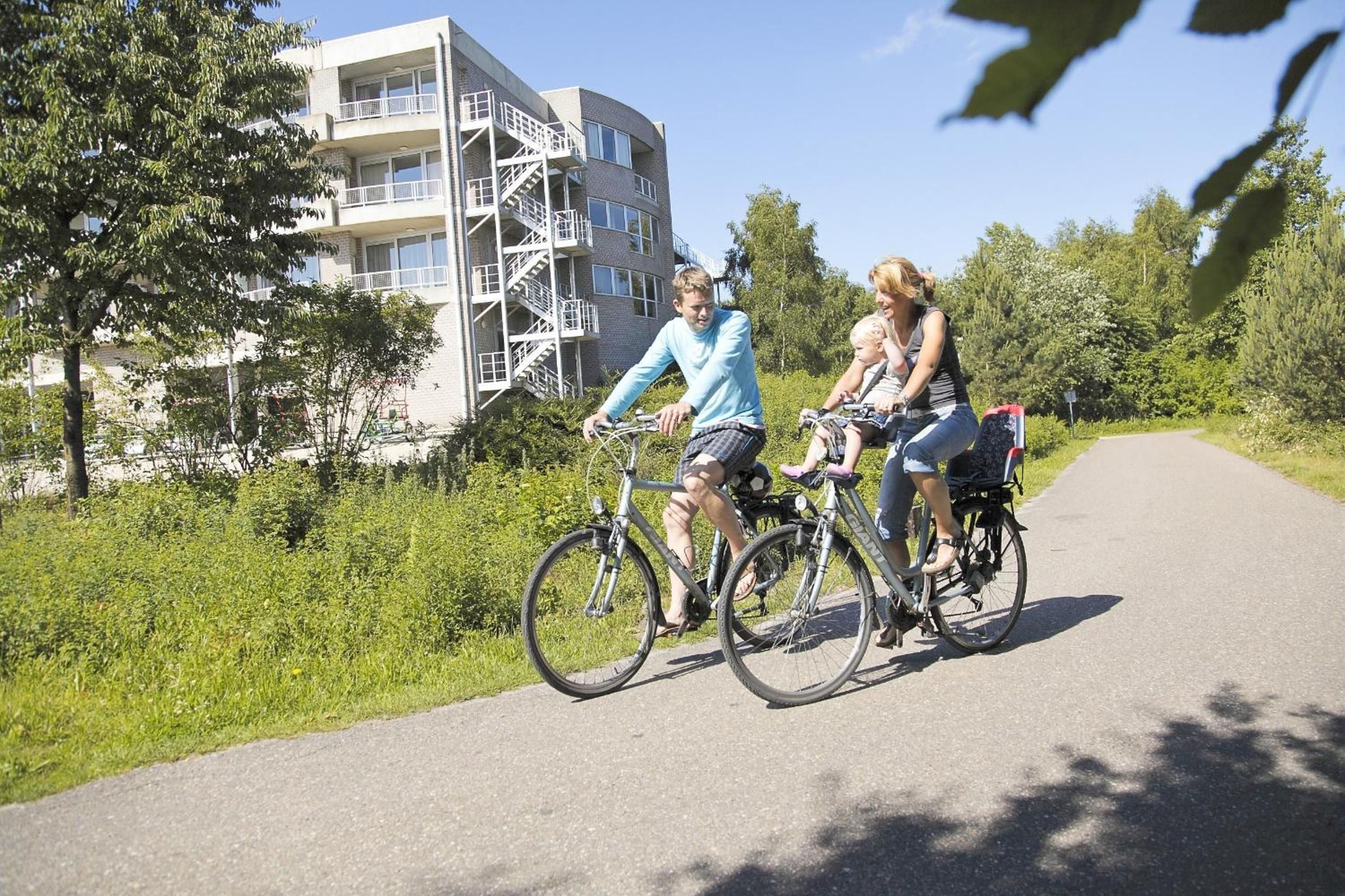 Un chemin pavé, fréquenté par des cyclistes, bordé de verdure et avec un immeuble à plusieurs étages en arrière-plan.