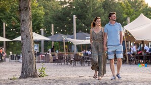 Una pareja caminando sobre una playa arenosa con árboles y sombrillas al fondo.