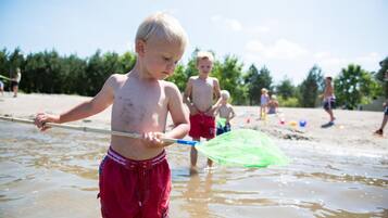 Un enfant joue dans une zone peu profonde près du rivage, sur une plage, tandis que d’autres enfants et des adultes se trouvent à l’arrière-plan.