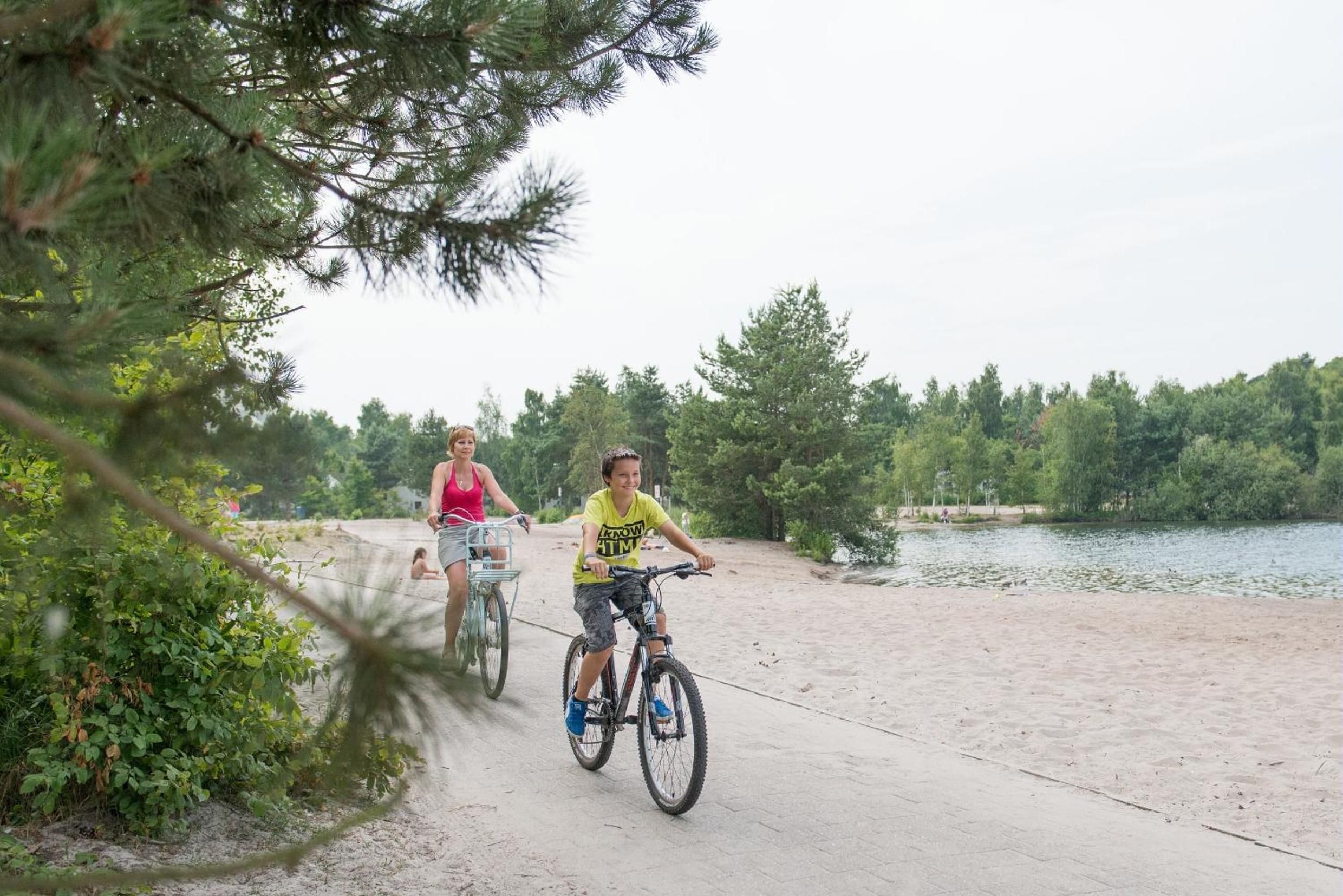 Two people riding bicycles on a concrete path by a lake surrounded by trees.